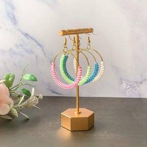 Può includere: Esposizione di orecchini a cerchio fatti a mano in vari colori, tra cui rosa, verde, blu e bianco. Gli orecchini sono appesi a un supporto color oro. I cerchi sono adornati con un design macramè, che aggiunge consistenza e interesse visivo ai gioielli.