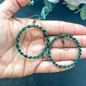 Può includere: Un paio di orecchini a cerchio in macramè verde fatti a mano. Gli orecchini presentano un design a spirale e sono sospesi da ganci dorati. Gli orecchini sono tenuti in una mano.