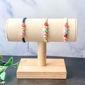 May include: Three beaded bracelets with colourful letter beads spelling out names. The bracelets are displayed on a beige jewellery stand with a wooden base. One bracelet is navy blue, one is light green, and one is white.
