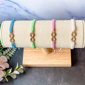 May include: Four macramé bracelets in blue, green, pink, and white, each with a gold-coloured infinity charm. The bracelets are displayed on a beige cylindrical stand with a wooden base, showcasing the jewellery's design and colours.
