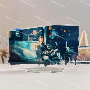 May include: A clear acrylic desk ornament featuring a watercolor illustration of a person seated, surrounded by planets and scientific diagrams. The artwork has a space and science theme, with a blue and yellow color palette.