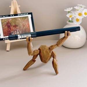 May include: A dark blue pen held by a brown wooden articulated figure. The figure is posed as if lifting the pen. In the background, a small framed picture on an easel displays the text "HAIL MARY". A white vase with daisies is also visible.
