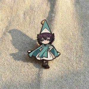 May include: A metal enamel pin featuring a cartoon character with dark purple hair, wearing a teal pointed hat with a small yellow crescent moon, a matching teal cape, and a white dress. The character has dark brown shoes and a serious expression.