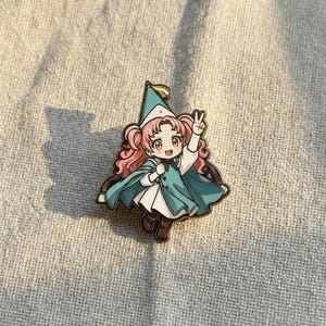 May include: A metal enamel pin featuring a cartoon character with pink pigtails, wearing a teal cape, a matching pointed hat, and a white dress. The character is making a peace sign with one hand and has a cheerful expression with large eyes.