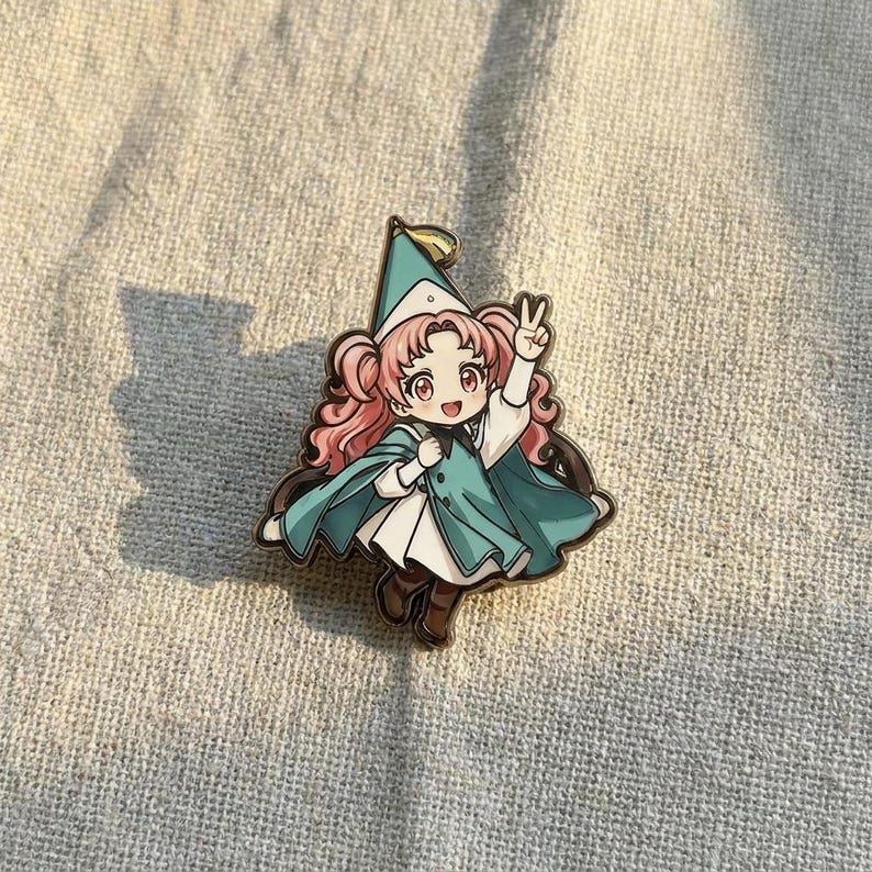 May include: A metal enamel pin featuring a cartoon character with pink pigtails, wearing a teal cape, a matching pointed hat, and a white dress. The character is making a peace sign with one hand and has a cheerful expression with large eyes.