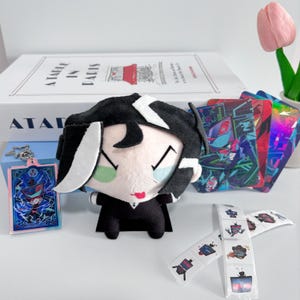 May include: A plush doll with black hair and white streaks, wearing a black suit, accompanied by a character keychain, three colourful rectangular cards with digital art, and a strip of small square stickers featuring television-headed characters.