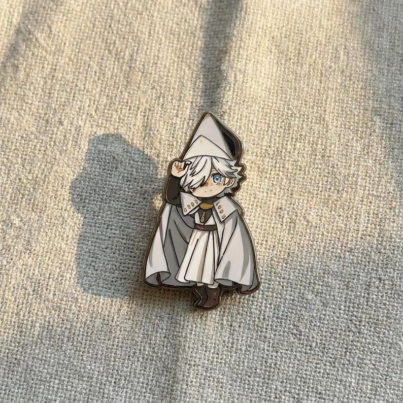 May include: A metal enamel pin featuring a stylized anime-style character with white hair and blue eyes. The character wears a white pointed hat, a white cape with dark grey lining, and a white tunic with gold-colored accents and a brown belt.