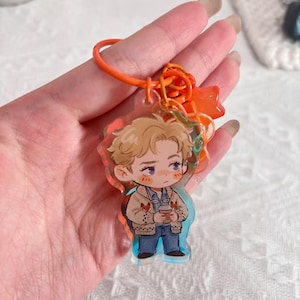 May include: An acrylic keychain featuring a cartoon character with blonde hair, wearing a beige cardigan with small fox patterns over a blue shirt and jeans, holding a coffee cup. The charm is attached to a bright orange ring and a star-shaped charm.