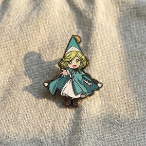 May include: A metal enamel pin featuring a cartoon character with short blonde hair, wearing a pointed teal hat with a yellow tassel, a teal cape over a white dress, and dark brown boots. The character has large eyes and is reaching forward.