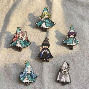 May include: Six enamel pins featuring chibi-style characters wearing pointed hats and capes. The characters have distinct hair colors including blonde, pink, dark purple, black, light blue, and white, each dressed in layered outfits with capes.