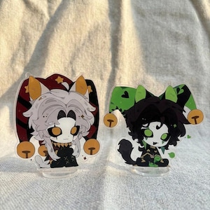 May include: Two acrylic character standees featuring stylized figures with jester hats. The left figure has white hair, orange eyes, and a red and black hat. The right figure has black hair, green eyes, and a green and black hat with bells.