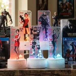 Könnte beinhalten: Eine Sammlung von LED-Nachtlichtern mit Resident Evil-Charakteren. Jedes Licht zeigt ein Charakterbild mit dem Wort "RESIDENT" vertikal aufgedruckt. Die Sockel haben einen Rissglaseffekt und strahlen sanft. Die Lichter werden mit Videospielboxen ausgestellt.