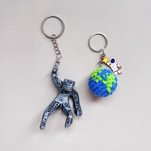 May include: Two keychains with silver-tone rings. One features a black and white abstract figure. The other has a blue and green crocheted globe with a small astronaut and star charm. The keychains are on a light background.