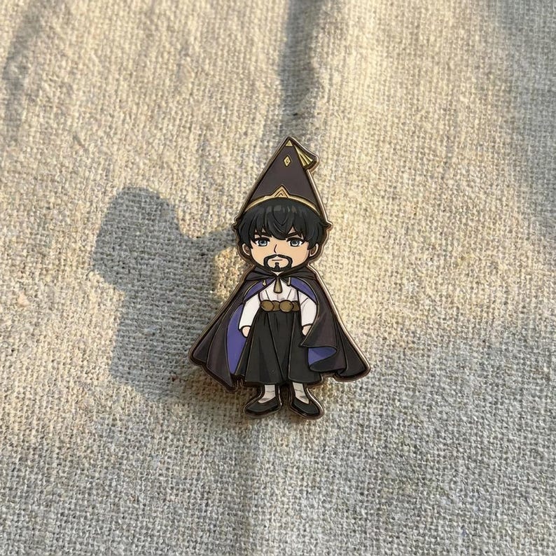 May include: A metal enamel pin featuring a stylized cartoon character with black hair and a beard, wearing a pointed hat, a white shirt, a dark skirt, and a long cape with a purple lining. The character has a gold-colored belt and trim.