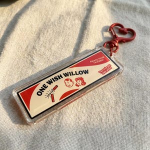 Puede incluir: Un llavero rectangular con un cierre rojo en forma de corazón. El llavero presenta el texto "ONE WISH WILLOW" con ilustraciones de un niño y una niña, y un petardo. El texto adicional dice "AMAZE YOUR FRIENDS!" y "You only get ONE WISH."