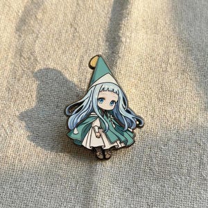May include: A metal enamel pin featuring a cartoon character with long light blue hair, wearing a pointed teal hat and a matching teal cape over a white dress. The character has large blue eyes and is depicted in a stylized, illustrated form.