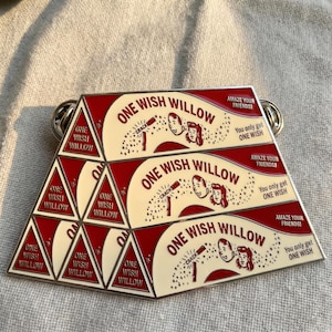 May include: A pyramid-shaped pin with a red and cream color scheme. The pin features the text "ONE WISH WILLOW" and illustrations of a couple and a magic wand. The text "AMAZE YOUR FRIENDS!" and "You only get ONE WISH" are also included.