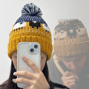 May include: A mustard yellow knit beanie featuring a repeating pattern of white and black sheep around the center. The hat has a ribbed cuff, a dark blue and white patterned top section, and a matching dark blue and white pom-pom on the crown.