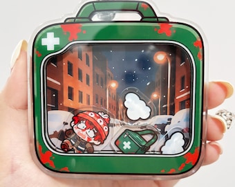 May include: A clear acrylic shaker keychain shaped like a green first aid kit with red blood splatters. Inside, a cartoon character with red hair wearing a beanie and a green medical bag move against a snowy city street background.