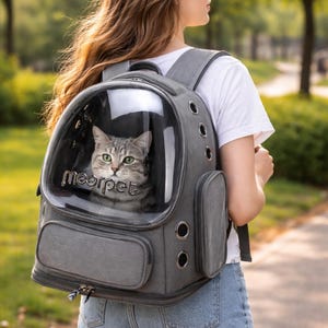 Puede incluir: Mochila transportadora gris para gatos con ventana de burbuja transparente. La mochila tiene un exterior gris con múltiples orificios de ventilación y un bolsillo lateral. El gato dentro tiene ojos verdes y está mirando hacia afuera. El texto "meowpet" es visible.