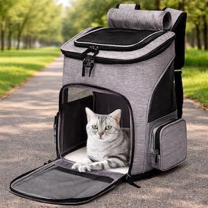 Puede incluir: Una mochila transportadora gris para gatos con un gato dentro, sentado sobre una almohadilla blanca. El transportador tiene una puerta frontal de malla y un bolsillo lateral. El gato es gris con ojos verdes.