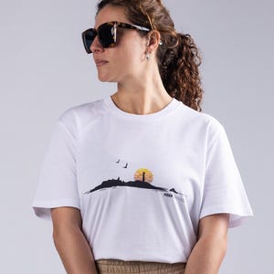 Puede incluir: Camiseta blanca con un gráfico de paisaje que presenta un sol amarillo, una silueta negra de una colina y pájaros. La persona lleva pantalones color caqui, gafas de sol negras, un bolso beige y zapatos negros estilo alpargata.