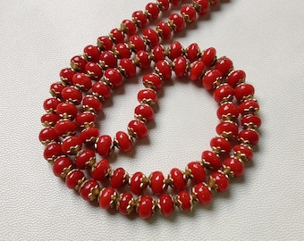 Puede incluir: Un collar vintage con cuentas rojas vibrantes y detalles dorados. Las cuentas ovaladas están ensartadas, creando un efecto en capas. El collar se muestra sobre un fondo neutro.