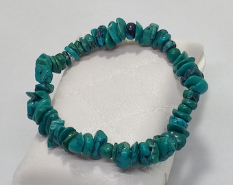 Può includere: Un braccialetto di perline di turchese. Il braccialetto è composto da pietre di turchese irregolari e lucide in varie tonalità di blu-verde. Una singola perla scura è intercalata tra le schegge di turchese. Il braccialetto è esposto su una superficie bianca.