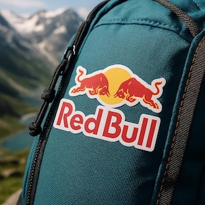 Könnte beinhalten: Türkisfarbener Rucksack mit einem Red Bull Logo-Aufnäher. Der Aufnäher zeigt zwei rote Bullen vor einer gelben Sonne, mit dem Markennamen in roten Buchstaben. Der Rucksack hat schwarze Reißverschlüsse und Riemen, mit einer Berglandschaft im Hintergrund.