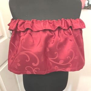 Puede incluir: Un top rojo sin tirantes con un escote con volantes. La tela tiene un sutil estampado floral en relieve. El top se muestra en un maniquí negro, mostrando su diseño y ajuste.