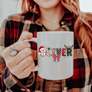 K&ouml;nnte beinhalten: Wei&szlig;e Keramik-Tasse mit dem Namen "OLIVER" in einem festlichen Weihnachtsdesign. Die Buchstaben sind mit einem Weihnachtsmannhut, einer Schleife und Rentieren verziert, mit Schneeflocken um den Namen. Ein perfektes Weihnachtsgeschenk.