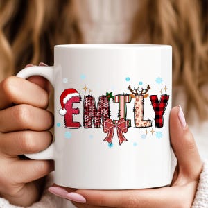 K&ouml;nnte beinhalten: Wei&szlig;e Keramik-Tasse mit dem Namen "EMILY" in einem weihnachtlichen Design. Die Buchstaben sind mit Weihnachtsm&uuml;tzen, Schneeflocken, Karomustern und Rentieren verziert. Eine rote Schleife und Stechpalmenbl&auml;tter vervollst&auml;ndigen das festliche Thema.