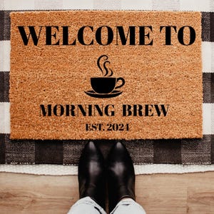 May include: A rectangular doormat with the words "WELCOME TO MORNING BREW EST. 2024" in black lettering. A black coffee cup graphic is centered above the words. The mat is made of a natural fiber material and is placed on a black and white checkered rug.