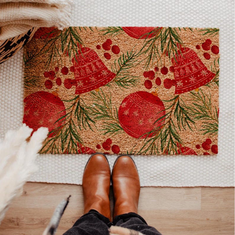 May include: A rectangular doormat with a festive Christmas design. The mat features a repeating pattern of red bells, ornaments, and berries, along with green pine needles, set against a tan background. The mat is made of a natural fiber material.