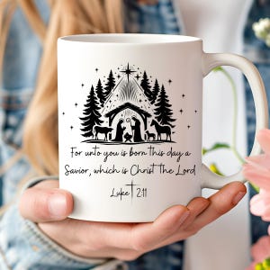 Könnte beinhalten: Weißer Keramikbecher mit einer schwarzen Silhouette einer Krippenszene, Bäumen und Sternen. Der Text "For unto you is born this day a Savior, which is Christ the Lord Luke 2:11" ist darunter gedruckt. Der Becher hat einen weißen Henkel.
