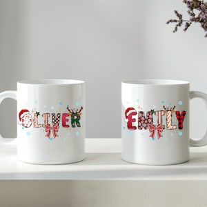 K&ouml;nnte beinhalten: Zwei wei&szlig;e Keramikbecher mit den Namen "Oliver" und "Emily" in festlicher Schrift. Die Namen sind mit Weihnachtselementen wie Weihnachtsm&uuml;tzen, Rentiergeweihen und Schleifen verziert. Die Becher stehen auf einer wei&szlig;en Oberfl&auml;che.