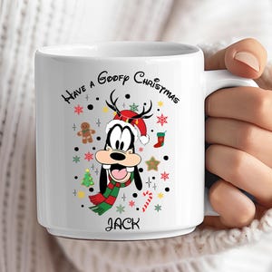 Könnte beinhalten: Weiße Keramik-Tasse mit einem weihnachtlichen Goofy-Design. Die Tasse trägt den Text "Have a Goofy Christmas" und den Namen "Jack". Das Design zeigt Goofy mit Weihnachtsmütze und Schal, umgeben von Weihnachtsdekorationen.