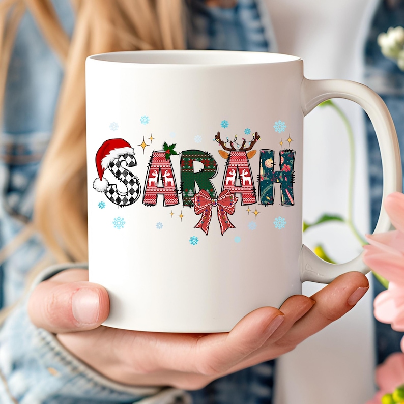 K&ouml;nnte beinhalten: Wei&szlig;e Keramik-Tasse mit dem Namen "SARAH" in festlichem Design. Die Buchstaben sind mit Mustern verziert, darunter ein Weihnachtsmannhut, Rentiergeweih und eine rote Schleife. Schneeflocken und Sterne akzentuieren das Design.