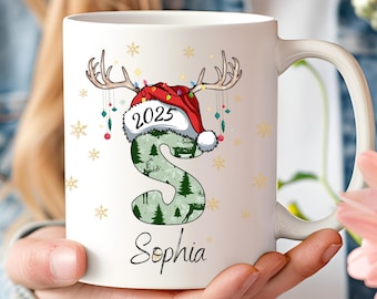 Op de afbeelding: Witte keramische mok met een kerstthema. De letter "S" is versierd met een bospatroon en een kerstmuts met geweien. Het jaar "2025" staat op de muts en de naam "Sophia" staat eronder.