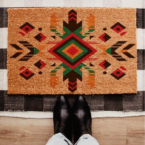 May include: A rectangular doormat with a geometric design in shades of red, green, orange, and brown. The design is centered on a natural coir background. Black boots and blue jeans are visible at the bottom of the image.