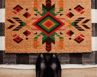 May include: A rectangular doormat with a geometric design in shades of red, green, orange, and brown. The design is centered on a natural coir background. Black boots and blue jeans are visible at the bottom of the image.