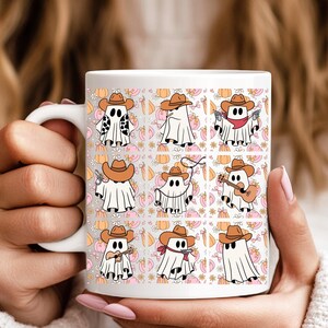Könnte beinhalten: Weiße Keramik-Tasse mit einem sich wiederholenden Muster von Cartoon-Geistern mit Cowboyhüten, Bandanas und verschiedenen Gegenständen. Der Hintergrund zeigt Kürbisse und florale Elemente in Orange, Rosa und Beige.