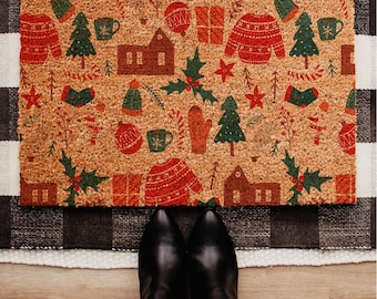 May include: A rectangular doormat with a festive holiday pattern. The mat features a tan background with repeating images of red sweaters, green Christmas trees, houses, and other holiday-themed elements. Black boots are visible at the bottom of the image.
