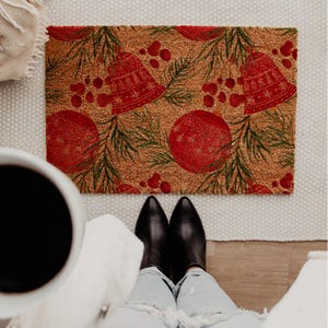 May include: A rectangular doormat with a festive Christmas design. The mat features red bells, ornaments, and berries, along with green pine needles, set against a brown background. The mat is made of a durable material.