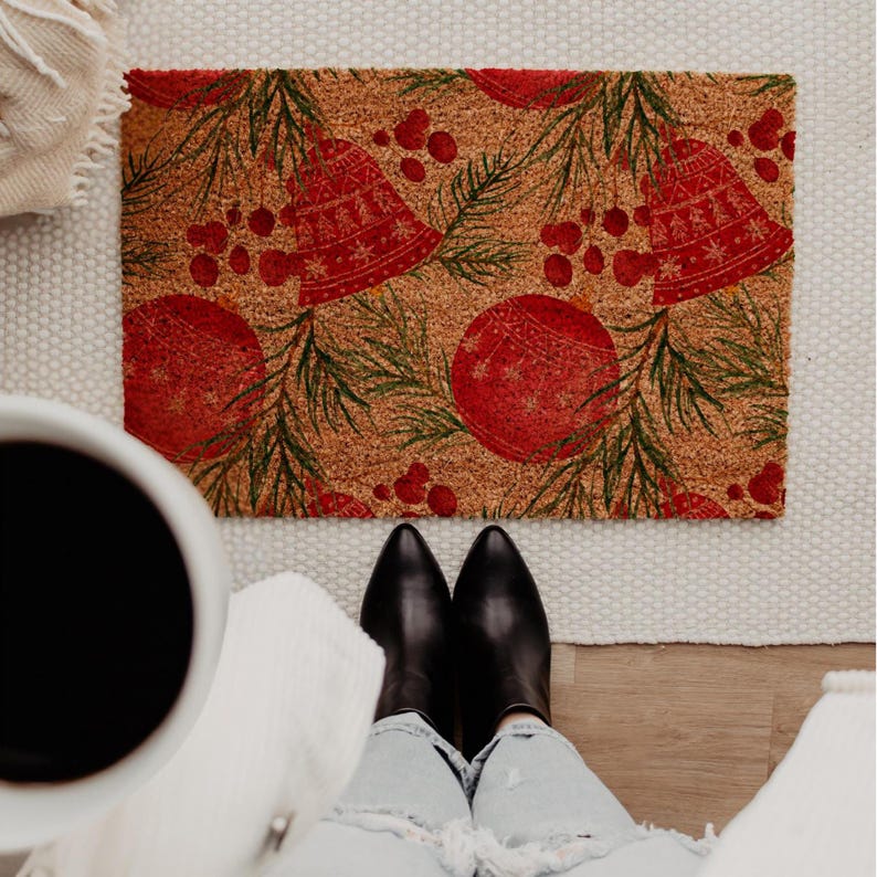 May include: A rectangular doormat with a festive Christmas design. The mat features red bells, ornaments, and berries, along with green pine needles, set against a brown background. The mat is made of a durable material.