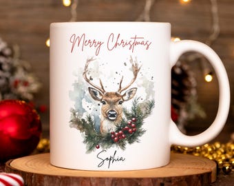 May include: White ceramic mug with a watercolor illustration of a deer, pine branches, and red berries. The words "Merry Christmas" and a name are written in red script. The mug is on a wooden coaster.