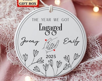 Könnte beinhalten: Weißes Keramik-Verlobungsornament mit dem Text "The Year We Got Engaged", den Namen "Jeremy" und "Emily" und dem Jahr "2025". Das Design umfasst eine Strichzeichnung von Händen mit einem Ring und floralen Details. Ein roter "Free Gift Box" Aufkleber befindet sich oben links.