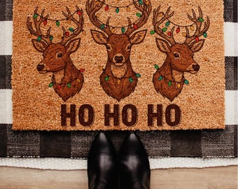 May include: A rectangular doormat featuring three brown reindeer heads decorated with festive Christmas lights. The words "HO HO HO" are printed in brown below the reindeer. The mat is made of a natural fibre material.