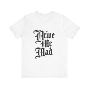 Puede incluir: Camiseta blanca con la frase "Drive Me Mad" impresa en fuente gótica. El texto está apilado verticalmente, con cada palabra en una línea separada. La camiseta está hecha de un material suave.