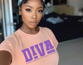 May include: A peach-colored t-shirt with the word "DIVA" in large purple letters. Below the word "DIVA" is the text "DIVINELY INSPIRED & VICTORIOUSLY ANOINTED" in smaller purple letters. The person is wearing a gold choker and a patterned headband.